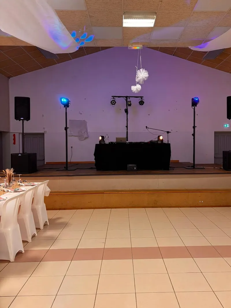 Scène d’une salle des fêtes prête pour la soirée : régie DJ, enceintes et projecteurs sur pieds, voilages au plafond